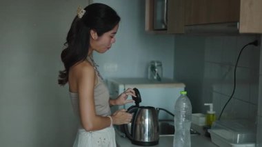 Genç bir kadın sabah kahvesi yapıyor, mutfak tezgahının yanında elektrikli çaydanlık dolduruyor.