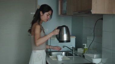 Asyalı kadın modern mutfakta kahvaltı hazırlıyor, bardağa sıcak su döküyor.