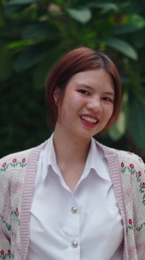 Beyaz gömlekli ve yün ceketli Asyalı genç kız parkta oturmuş gülümseyen kameraya bakıyor.