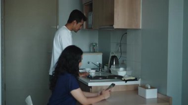 Genç çift birlikte yemek pişiriyor, kadın telefonunu karıştırıyor. Partnerin sebzeleri doğruyor. 4K Yatay