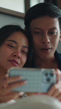 Gülümseyen genç çift yan yana uzanıyor, rahat yatak odası ortamında akıllı telefon ekranlarına bakıyorlar.