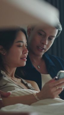 Gülümseyen Asyalı çift yatakta dinlenirken akıllı telefon ekranını kaydırıyor.