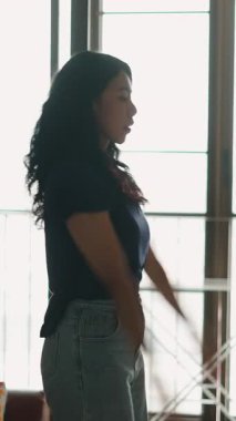 Geniş pencerenin yanında gevşeyen, esneyen, yumuşak doğal ışık alan kadın - 4K Yatay