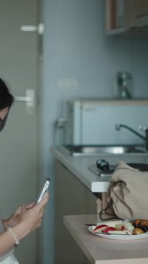 Genç iş kadını kahvaltı yapıyor, mermer mutfak tezgahının yanında akıllı telefon kaydırıyor.