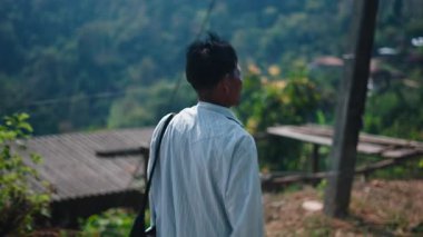 Asian farmer lahu walks along the dirt road with a fanny pack on his shoulder through a sprawling green valley - 4K Horizontal