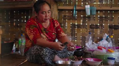 Asian woman peeling vegetables sitting on the floor cross-legged in a traditional kitchen with bamboo walls - 4K Horizontal