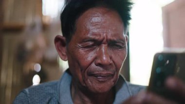 Close-up face and hands of Asian senior man focused on smartphone use inside a traditional lahu house in asia - 4K Horizontal