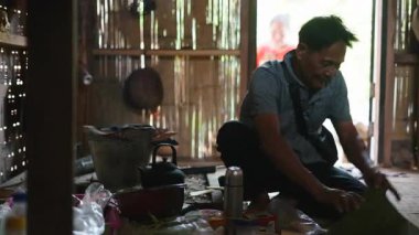 Southeast Asian man prepares and wraps traditional food inside banana leaves in a simple wooden kitchen - 4K Horizontal