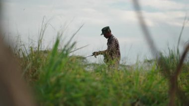 Asian Man in a camouflage cap and plaid shirt fishing with a rod surrounded by greenery in Ban Bang Phra - 4K horizontal