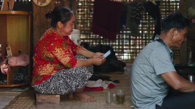 Asian woman from the lahu tribe sitting barefoot in her wooden hut consults her smartphone screen calmly while drinking hot tea - 4K Horizontal