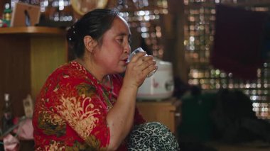 Lahu woman quietly drinking hot tea from a crystal glass in her traditional wooden and reed hut - 4K Horizontal