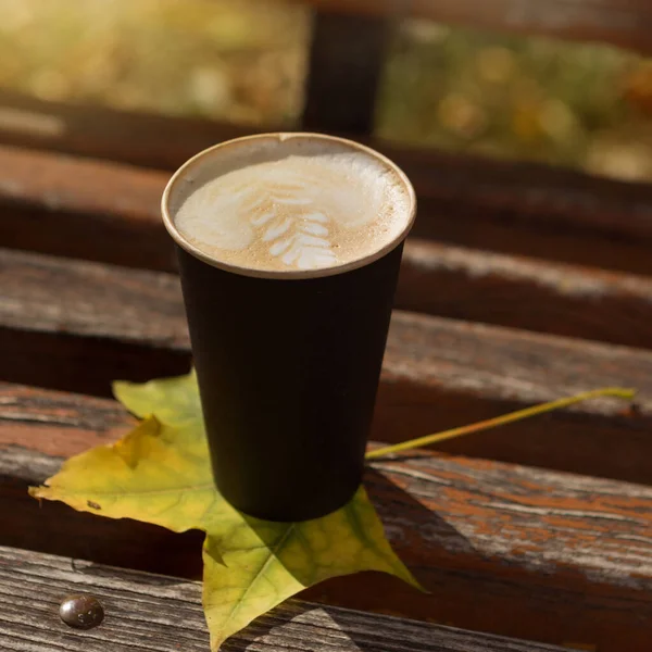 A cardboard glass with a cappuccino drink, stands on a bench, in an autumn park, close-up