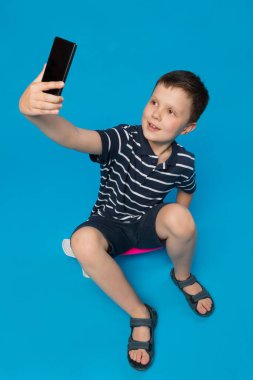 Neşeli çocuk kaykayın üzerinde oturur ve akıllı bir telefonla elini kaldırır. Mavi arka planda selfie, konsept çeker.
