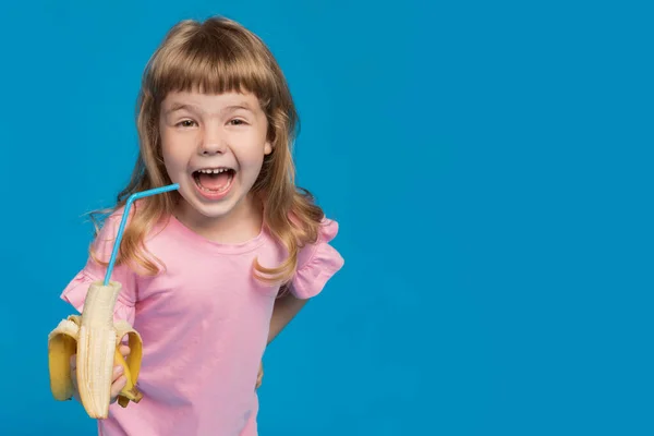 ピンクのブラウスに身を包んだ少女が手に持っているバナナにカクテルチューブと笑い感情とジェスチャー夏と健康的な食事の概念青い背景にコピースペース — ストック写真