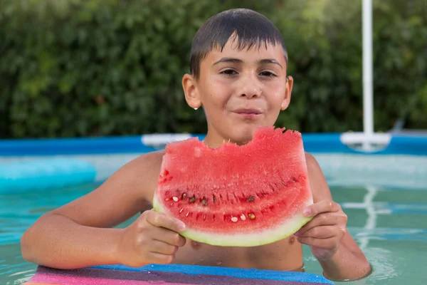 wet boy in the pool holds a large piece of watermelon in his hands and eats it, looks at the camera