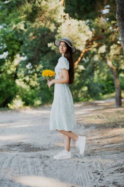 Şapkalı güzel bir kız ve elinde bir buket çiçekle ormanda yürüyor..