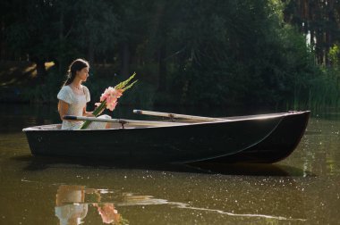 Güzel genç bir kız sabah erkenden elinde bir buket çiçekle bir orman gölünde yelken açıyor.
