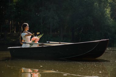 Güzel genç bir kız sabah erkenden elinde bir buket çiçekle bir orman gölünde yelken açıyor.
