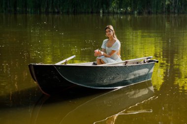 Güzel genç bir kız sabah erkenden elinde bir buket çiçekle bir orman gölünde yelken açıyor.