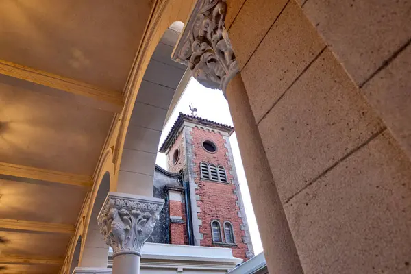 stock image View of bell tower of an abbey through an arch formed by Corinthian columns. At the top of the tower, there is a black rooster weather vane, contrasting with the sky