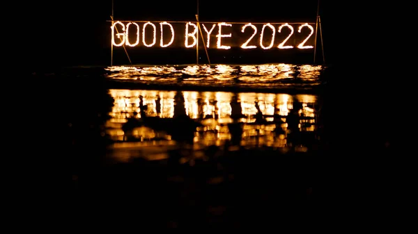 good bye 2022 burning phrase made of fire stick placed on water in sea, happy new year celebration in tropical place at night, inscription on the beach