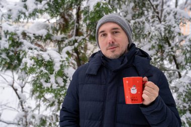 Gülümseyen adam lezzetli bir çay fincanı tutuyor, kırmızı Noel bardağında kahve içiyor, dışarıda, karlı çam ağacının yanında. Günlük kıyafetler, gri şapka ve kışlık ceket giyiyor. Soğukta nasıl ısınırım 
