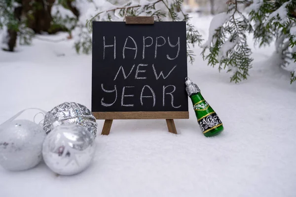 Happy new year wishes text inscription on blackboard. Silver balls baubles and champagne bottle toy decorations. Christmas greeting card stands on snow in forest. Festive winter holidays
