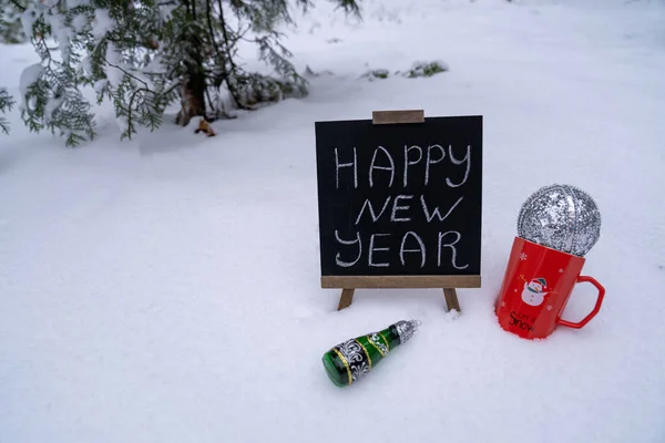 happy new year wishes text inscription on blackboard. Red mug with snowman print and silver ball standing on fresh snow in forest. New year toy decoration champagne. Festive winter holidays