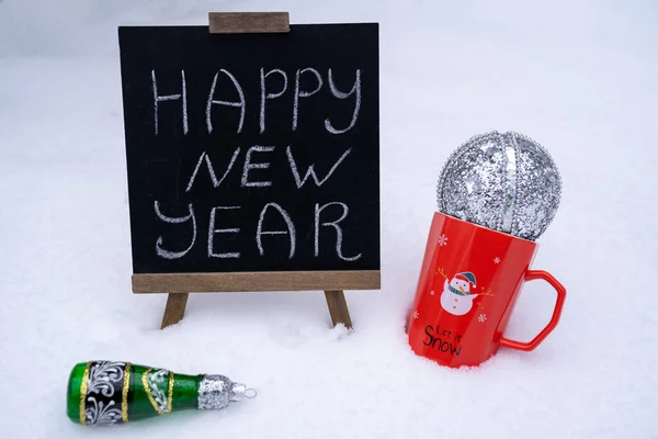 happy new year wishes text inscription on blackboard. Red mug with snowman print and silver ball standing on fresh snow. New year toy decoration champagne. Festive winter holidays