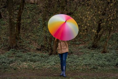Bej ceketli ve çizmeli genç bir kadın ormanda ya da parkta duruyor ve elinde renkli parlak gökkuşağı şemsiyesi tutuyor. Kız bulanık etki için şemsiye çeviriyor. Kapama hızı yavaş. Şehirde sonbahar havası