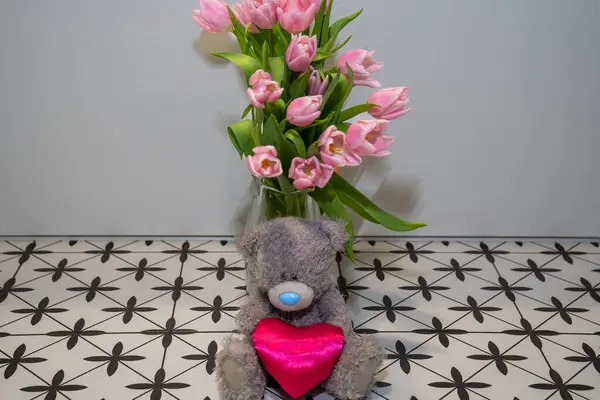 grey plush toy teddy bear with pink heart next to pink blooming flower bouquet of tulips in vase. Happy valentines day gift