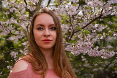 Pembe elbiseli güzel sarışın kadın çiçek açmış sakura ağacının yanındaki botanik bahçesi parkında poz veriyor. Bahar ve saflık, doğal güzellik. Uluslararası Kadınlar Günü