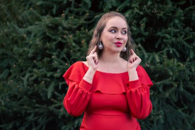 Yeşil bir köknar ağacının yanında duran kırmızı elbiseli mutlu genç bir kadın. Noel kutlaması kıyafeti. Tatil partisi.