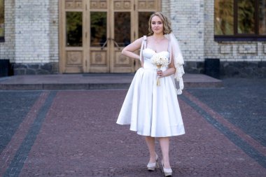 Beyaz kısa gelinlik ve duvak giymiş güzel genç gelinin tam boy portresi. Gelin elinde bir buket kurumuş çiçek tutuyor. Düğün günü.. 