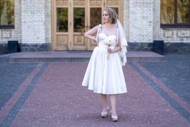 Beyaz kısa gelinlik ve duvak giymiş güzel genç gelinin tam boy portresi. Gelin elinde bir buket kurumuş çiçek tutuyor. Düğün günü.. 