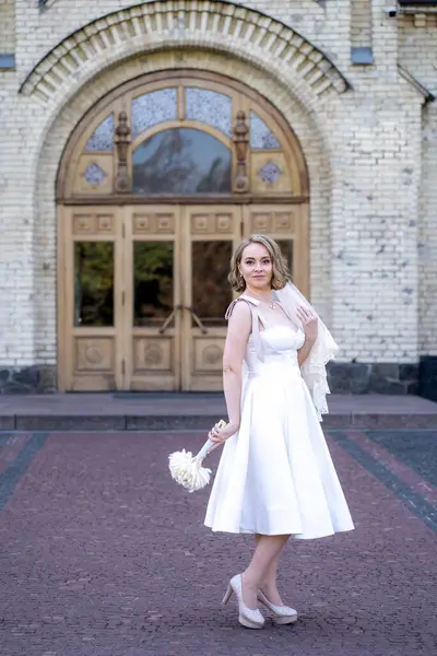 Beyaz kısa gelinlik ve duvak giymiş güzel genç gelinin tam boy portresi. Gelin elinde bir buket kurumuş çiçek tutuyor. Düğün günü.. 