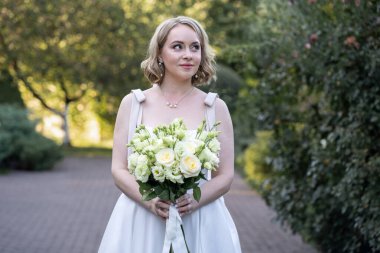 Beyaz çay uzunluğunda bir gelinlik içinde, elinde bir demet fildişi ve yeşil tomurcuklu allık güller olan zarif bir gelin, hafifçe yana bakıyor. Parke taşlı bir yolda romantik arka planda, ışık saçan zarafet ve güzellik dolu bir yolda yakalanmış..