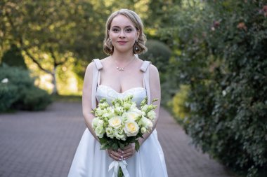 Beyaz çay uzunluğunda bir gelinlik giymiş, elinde bir demet fildişi ve yeşil tomurcuklu gül tutan bir gelinin portresi. Çimenlik, sonsuz zarafet ve dingin bir gelin eşliğiyle çevrili bir kaldırım taşından yolda yakalanmış.