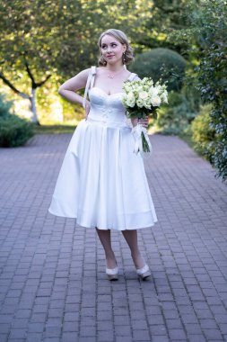 Beyaz gelinlik içinde, elinde bir buket fildişi ve allık gül tutan neşeli bir gelin büyüleyici bir ifadeyle eğlenceli bir poz veriyor. Romantik bir açık havada kaldırım taşı yolunda yakalandım. 
