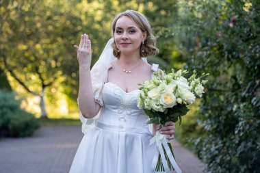 Beyaz bir gelinlik içinde elinde bir buket fildişi ve yeşil tomurcuklu kırmızı güller tutan mutlu gelin, gülümsüyor ve alyansını gösteriyor. Açık havada doğal ışık ve romantik yeşillik arka planlı bir düğün portresi.