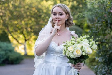 Beyaz gelinlik içinde gülümseyen gelin elinde bir buket fildişi ve yeşil tomurcuklu kırmızı güller, neşeli bir şşş hareketi yapıyor. Yumuşak doğal ışık ve yeşillik arka planlı romantik açık hava düğün portresi.