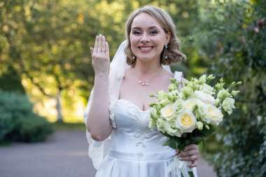 Beyaz bir gelinlik içinde elinde bir buket fildişi ve yeşil tomurcuklu kırmızı güller tutan mutlu gelin, gülümsüyor ve alyansını gösteriyor. Açık havada doğal ışık ve romantik yeşillik arka planlı bir düğün portresi.
