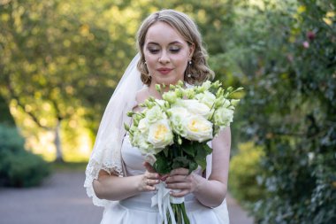 Beyaz gelinlikli gelin, elinde zarif bir demet fildişi ve yeşil tomurcuklu allık güller, çiçeklere nazikçe bakıyor. Yumuşak doğal ışık ve yeşillik arka planlı romantik açık hava düğün portresi.