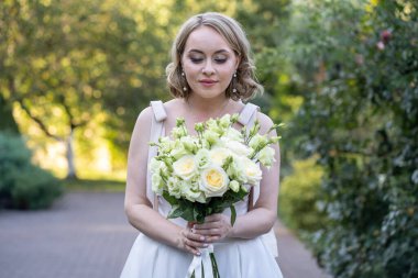 Beyaz gelinlikli gelin, elinde zarif bir demet fildişi ve yeşil tomurcuklu allık güller, çiçeklere nazikçe bakıyor. Yumuşak doğal ışık ve yeşillik arka planlı romantik açık hava düğün portresi.