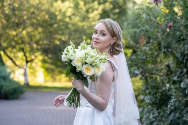 Beyaz gelinlik içinde güzel bir gelin elinde zarif bir demet fildişi ve narin yeşil tomurcuklu kırmızı güller tutuyor. Açık hava düğün portresi yumuşak doğal ışık ve yeşil arka plan, romantik ve kutlama temalı tasarımlar için mükemmel.