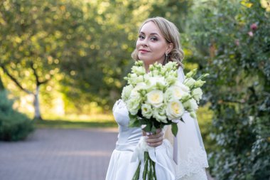 Beyaz gelinlik içinde güzel bir gelin elinde zarif bir demet fildişi ve narin yeşil tomurcuklu kırmızı güller tutuyor. Açık hava düğün portresi yumuşak doğal ışık ve yeşil arka plan, romantik ve kutlama temalı tasarımlar için mükemmel.