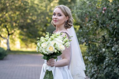 Beyaz gelinlik içinde güzel bir gelin elinde zarif bir demet fildişi ve narin yeşil tomurcuklu kırmızı güller tutuyor. Açık hava düğün portresi yumuşak doğal ışık ve yeşil arka plan, romantik ve kutlama temalı tasarımlar için mükemmel.