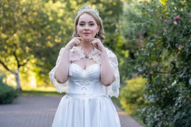 Beyaz saten elbiseli ve dantel pelerinli zarif gelin iki eliyle boynunu nazikçe tutuyor ve akşam ışığında dışarıda duruyor. Taç takıyor ve asil bir ifadesi var.