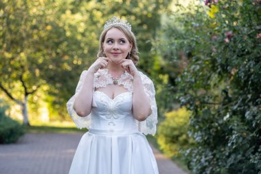 Çekici gelin beyaz bir gelinlik içinde dantelli bir pelerinle tatlı bir gülümsemeyle yan tarafa bakıyor. Tacı yumuşak bahçe ışığının altında parıldıyor.