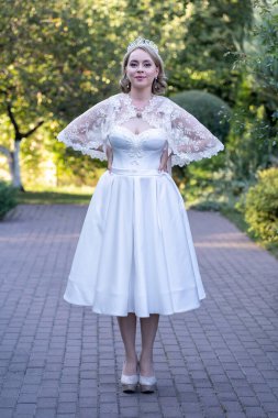 Kendine güvenen gelin elleri kalçasında, çay uzunluğunda beyaz bir gelinlik ve taç takmış dantelli bir pelerin giyiyor.